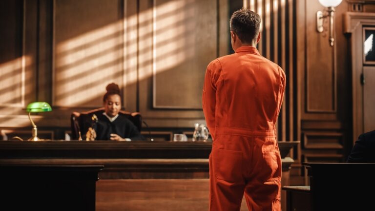 Man in orange prison jumpsuit standing before judge in wood-paneled courtroom.