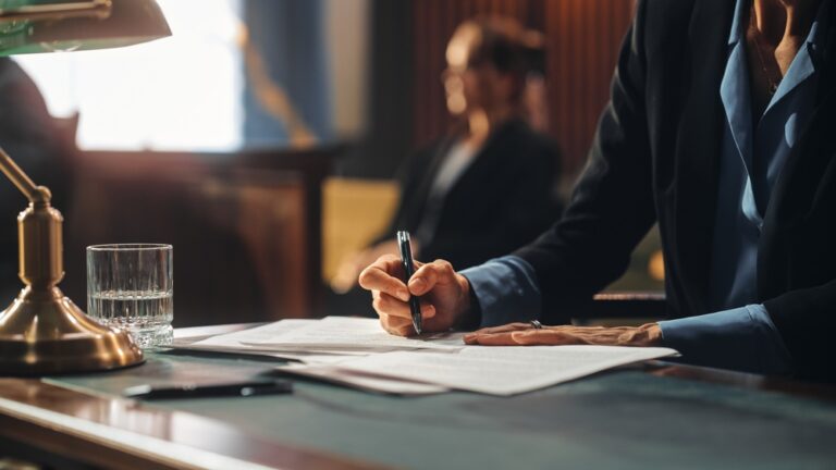 Person in dark suit writing on documents at desk with glass of water and lamp.