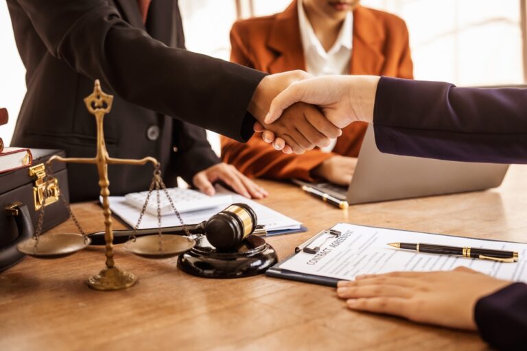 Two professionals shaking hands over a wooden desk with legal scales, gavel, and contract documents.