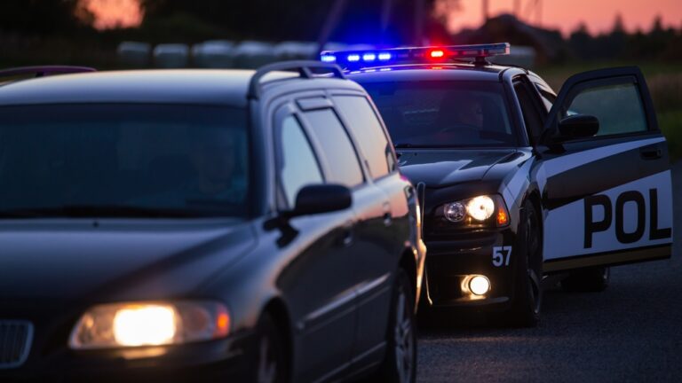 Police car with flashing lights and open door behind a stopped dark vehicle at dusk.