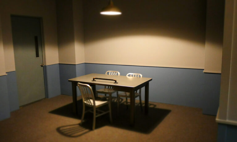 Empty interrogation room with metal table, three chairs, and overhead light.