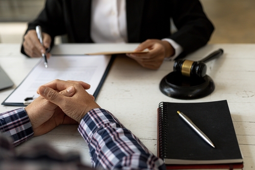 Two people seated at a desk with legal documents, a gavel, and a notebook with a pen.