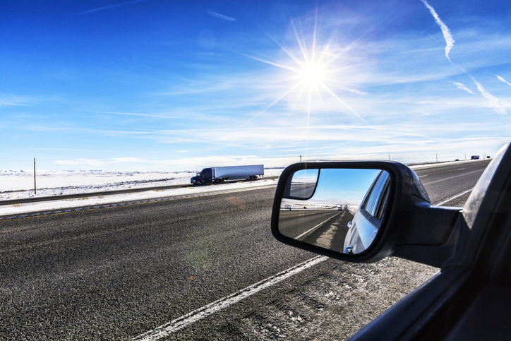 View of a highway with a semi-truck, bright sun, and a car's side mirror reflecting the road.