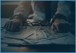 Person in handcuffs resting hands on table scattered with US dollar bills and eyeglasses.