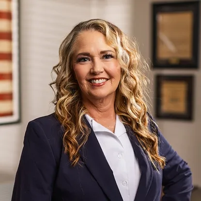 Professional portrait of a smiling woman with long blonde curly hair wearing a navy blazer and white blouse.
