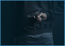 Person wearing dark clothing holding a black handgun behind their back.