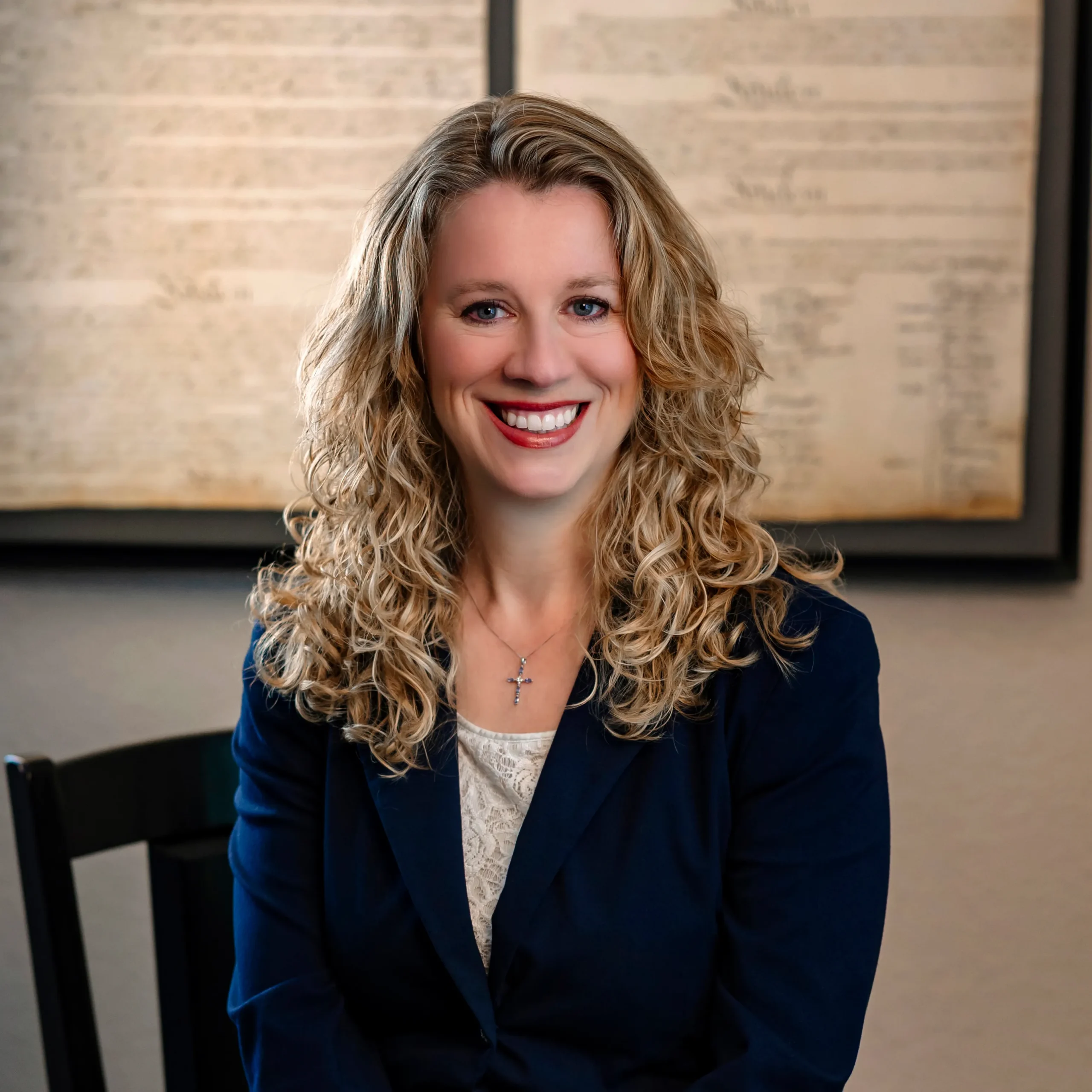 Professional portrait of a smiling woman with curly blonde hair and blue blazer, cross necklace visible.