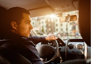 Man with short dark hair driving a car during sunset, interior dashboard visible.