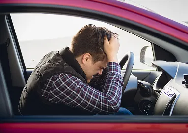Man in a red car holding his head in frustration, wearing a plaid shirt and black vest.