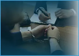 Two individuals with handcuffed hands resting on a table, one writing on a clipboard.