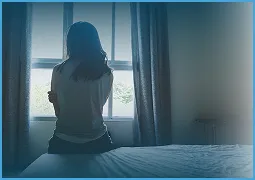 Woman in white shirt sitting on bed, looking out window with curtains.