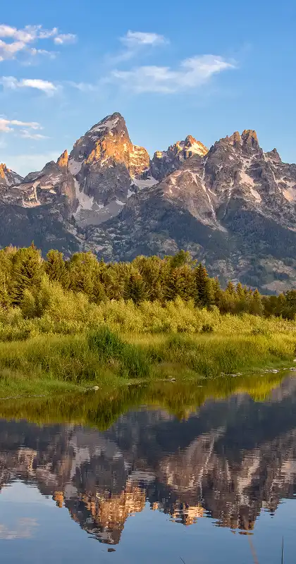 Sunlit rugged mountain peaks reflected in calm river with green forest foreground.