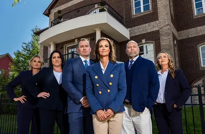 Group of six professionally dressed individuals standing confidently outdoors in front of a multi-story brick building.