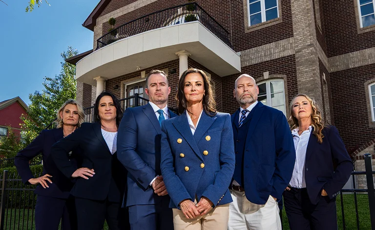 Group of six professionally dressed individuals standing confidently outside a brick building under a clear blue sky.