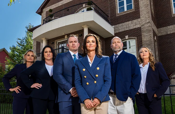 Group of six professionally dressed individuals standing confidently in front of a brick building.