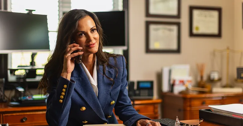 Professional woman in blue blazer speaking on phone in office with framed certificates.