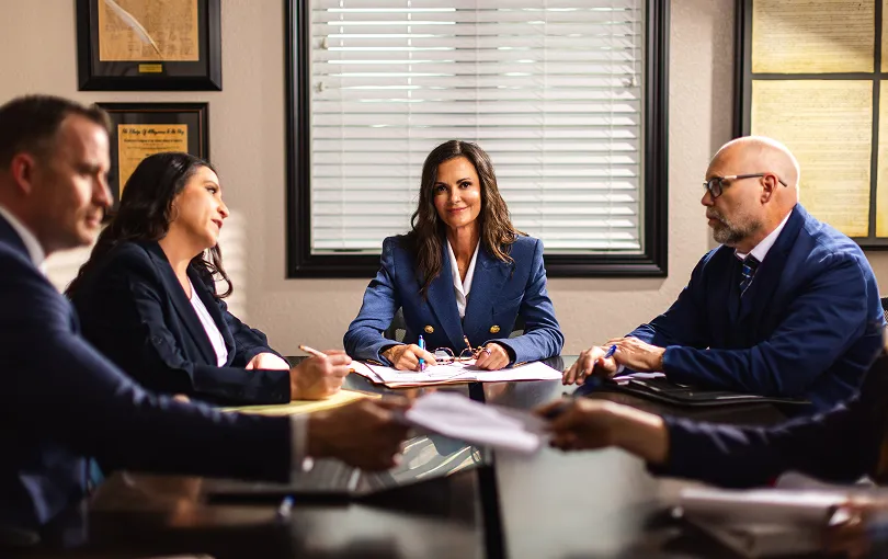 Professional business meeting with five individuals seated around a table, documents and pens visible.