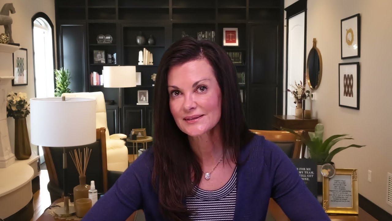 Professional woman with long dark hair seated in a well-lit room with dark bookshelves and beige walls.