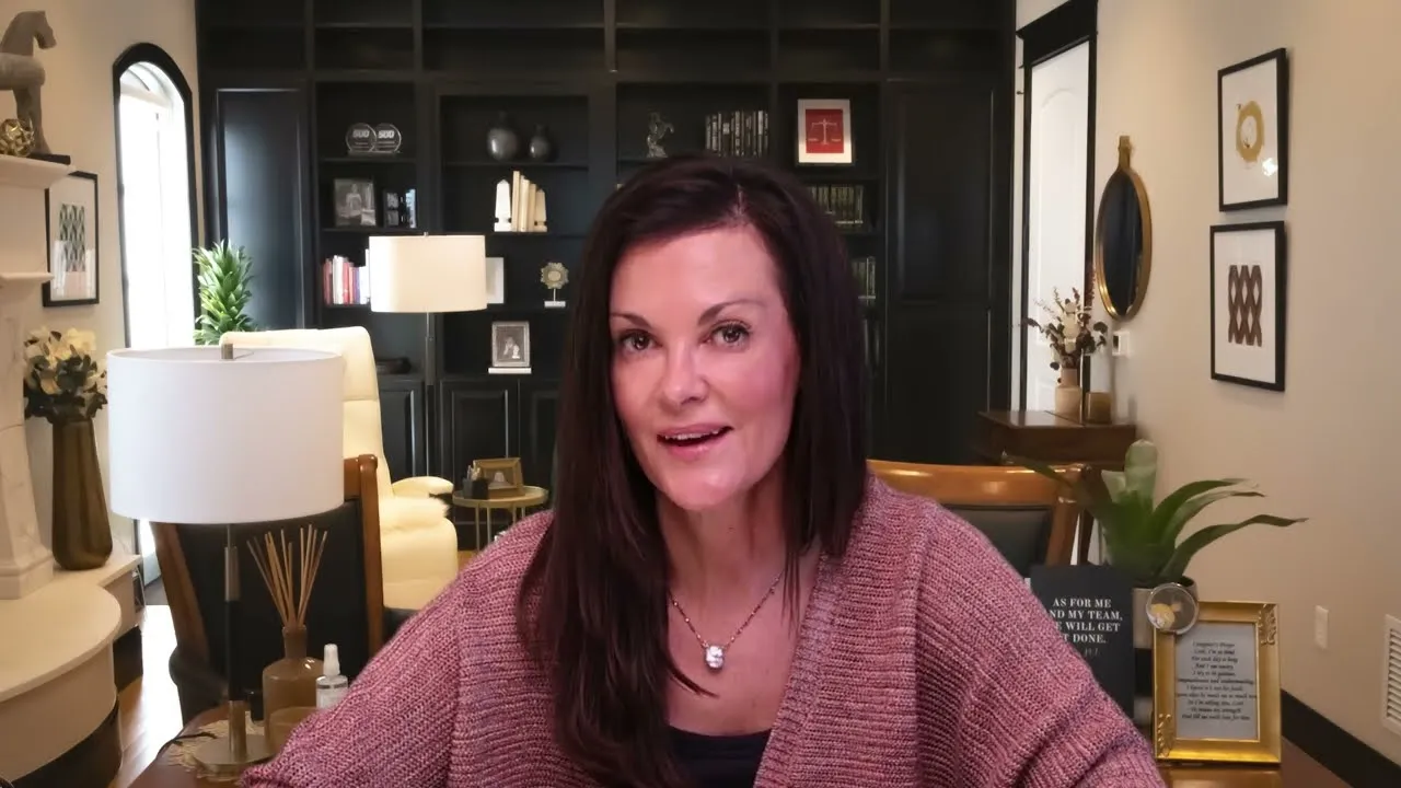 Woman with dark hair and pink sweater seated in a modern living room with black shelving and decorative items.