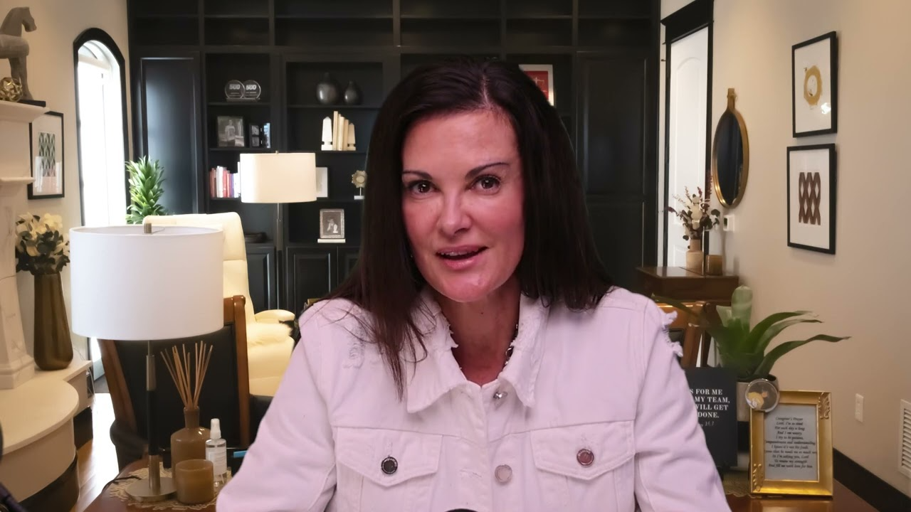 Middle-aged woman with dark hair wearing a white jacket seated in a modern living room with black shelves and decorative items.