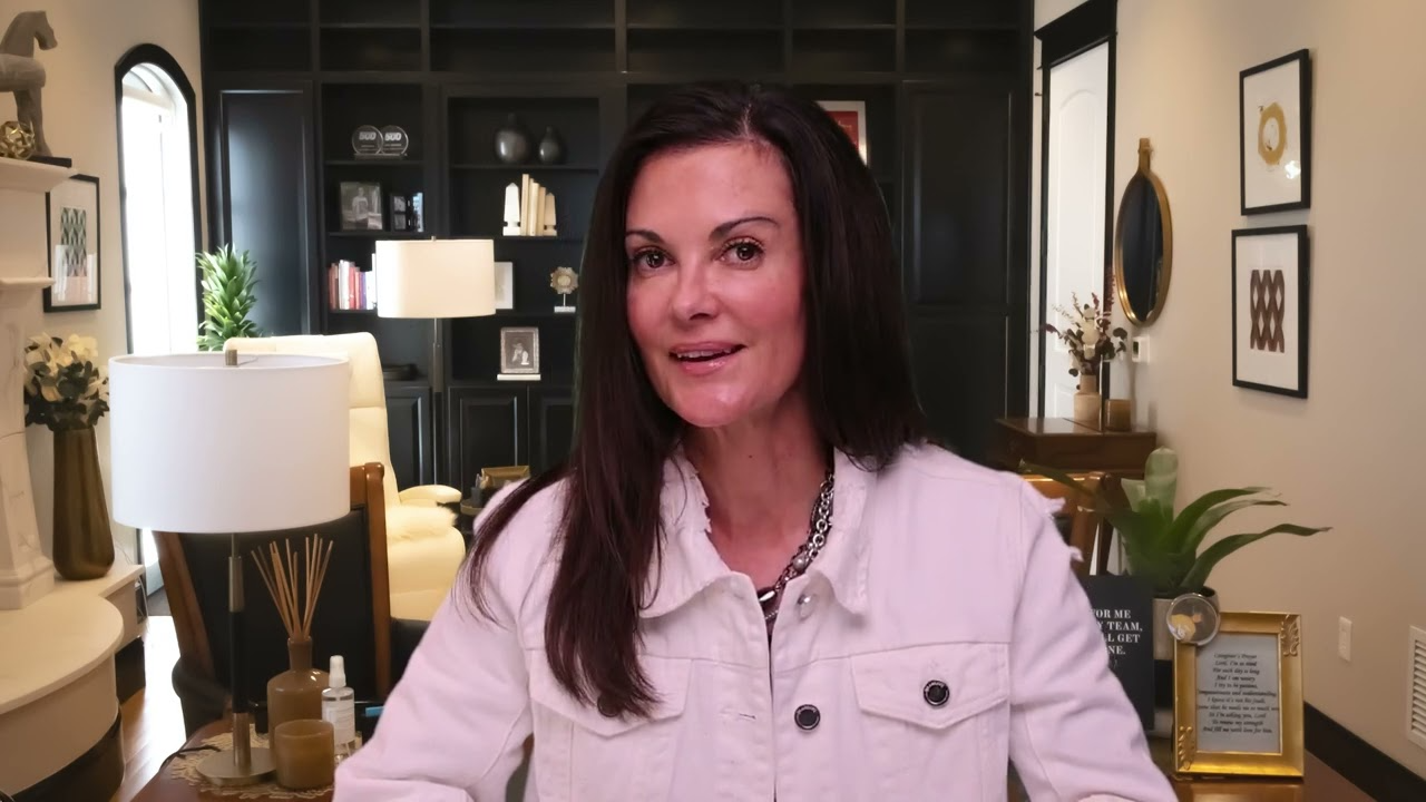 Woman with long dark hair wearing a white jacket seated in a modern, well-lit living room with dark shelves and decorative items.