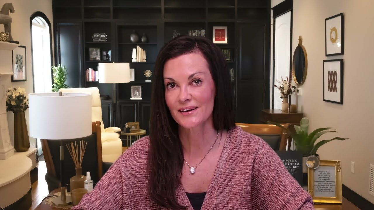 Professional woman with dark hair speaking in well-lit office with dark bookshelves and decorative plants.