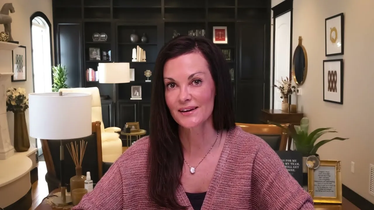 Woman with long dark hair wearing a mauve sweater seated in a well-lit, decorated living room.