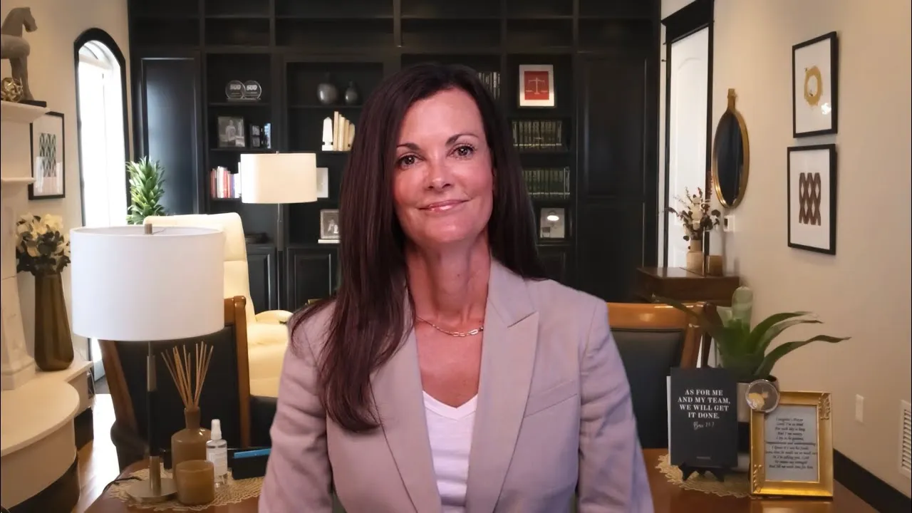 Professional woman with long brown hair wearing beige blazer, seated in modern office with dark shelves and decorative items.