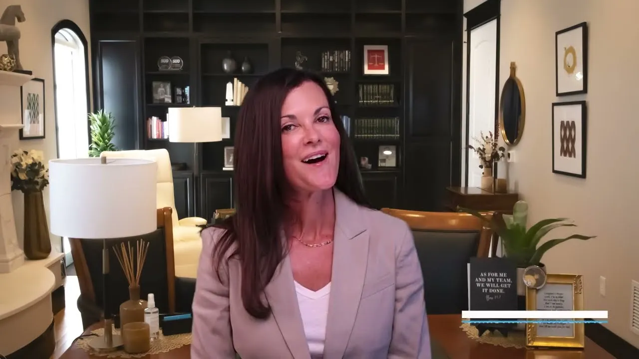 Professional woman with long dark hair in gray blazer speaking in a modern office with black bookshelves and decorative plants.
