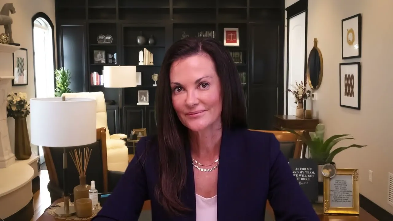 Professional woman with dark hair seated in a well-lit office with dark bookshelves and decorative items.