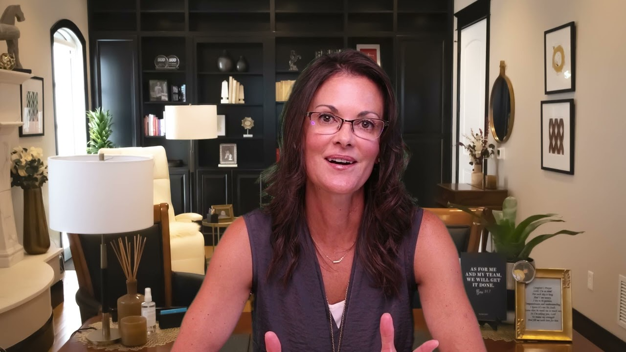Professional woman with glasses speaking in a modern office with dark bookshelves and decorative items.