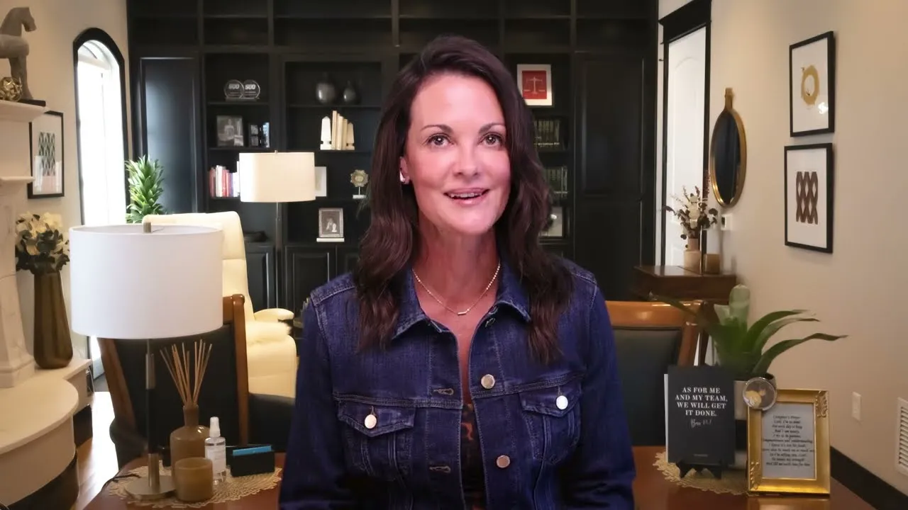 Woman with dark hair wearing a denim jacket seated in a warmly decorated living room with bookshelves and framed artwork.