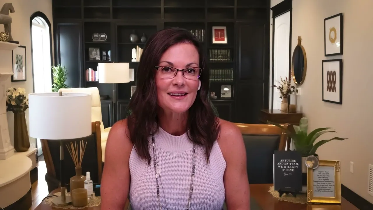 Middle-aged woman with glasses speaking, seated in a well-lit home office with dark bookshelves and decorative items.