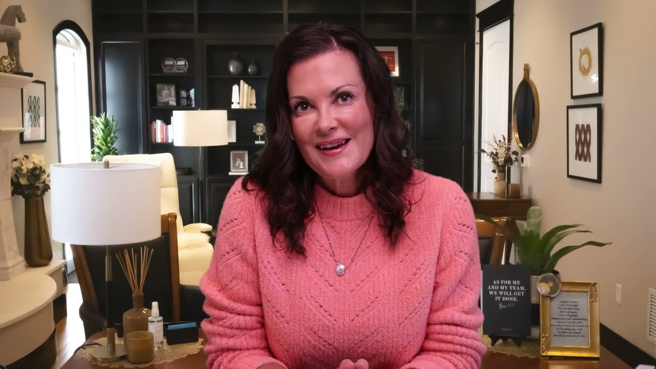 Woman in pink sweater sitting in a warmly decorated living room with dark shelves and framed quotes.