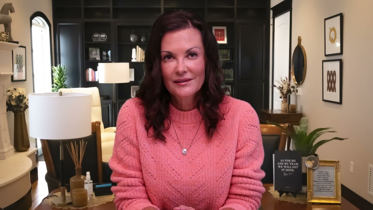 Woman in a pink sweater seated in a warmly lit room with bookshelves and framed art.