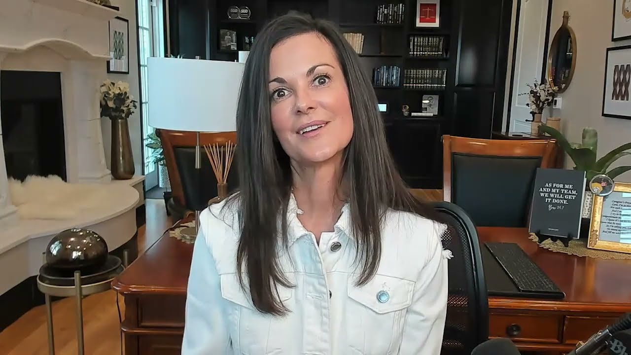 Professional woman with long brown hair wearing a white jacket, seated in an office with wooden furniture and bookshelves.