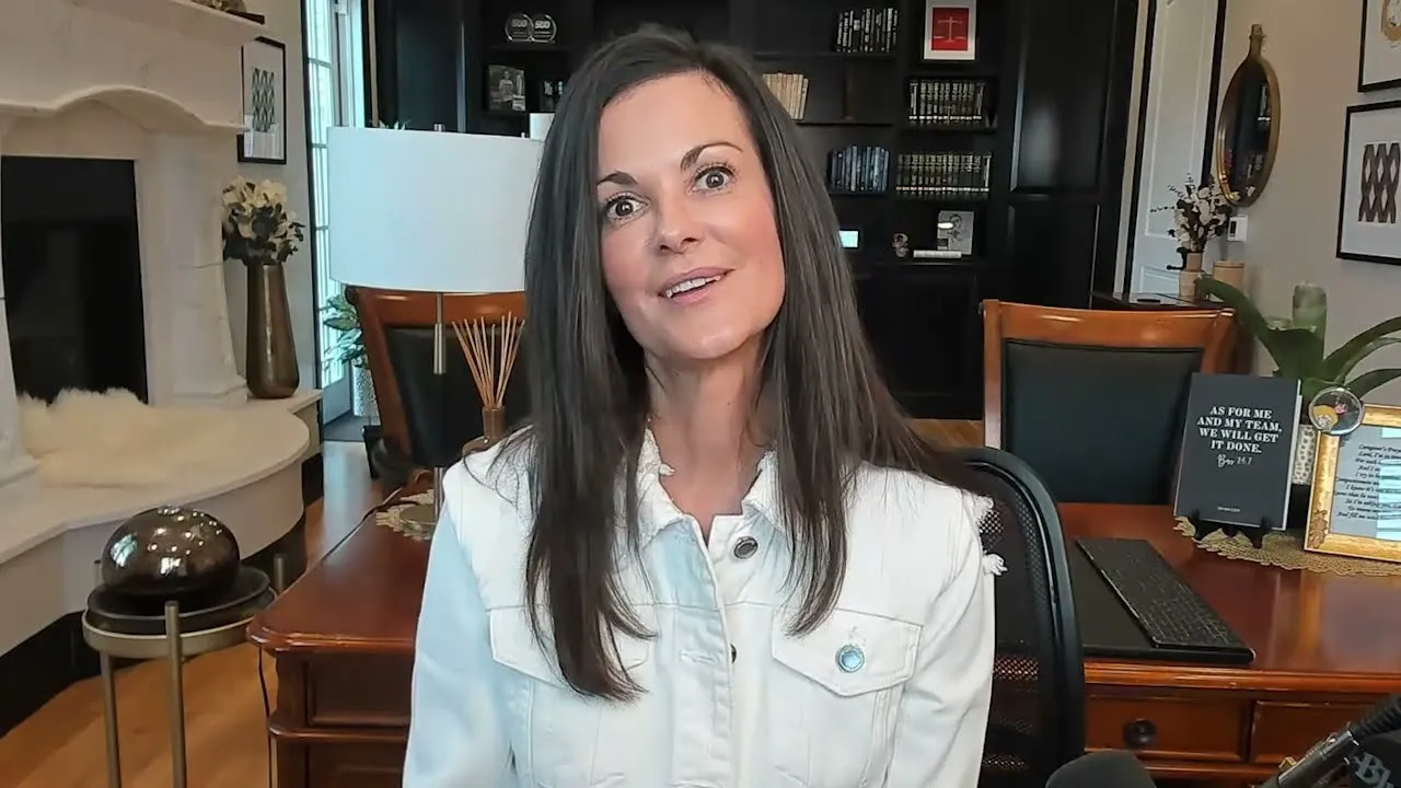 Professional woman with long dark hair wearing a white jacket, seated in a home office with wooden furniture and decor.