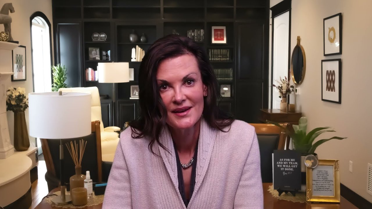 Professional woman with dark hair in a beige sweater seated in an elegant office with bookshelves and framed quotes.