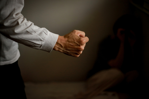Close-up of a clenched fist in foreground with a distressed person sitting in the dark background.