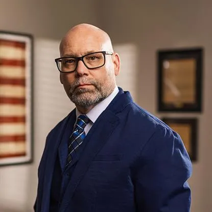 Portrait of a bald man with glasses wearing a navy suit and patterned tie in an office setting.