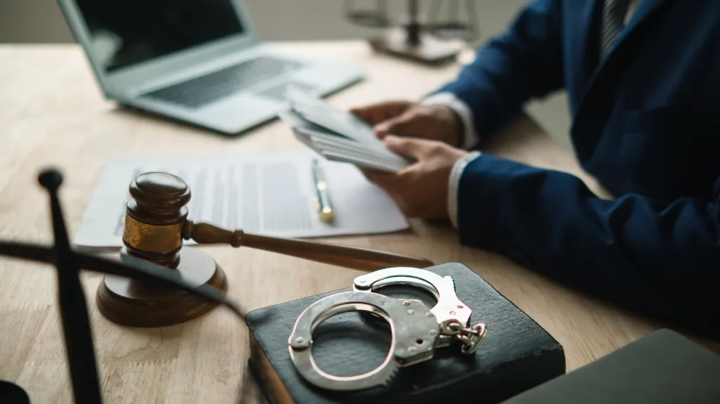 Close-up of handcuffs on a book, a judge's gavel, and a person in a suit reviewing documents.