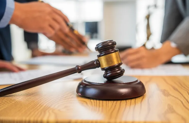 Wooden judge's gavel with brass band on a desk, blurred people discussing documents in background.