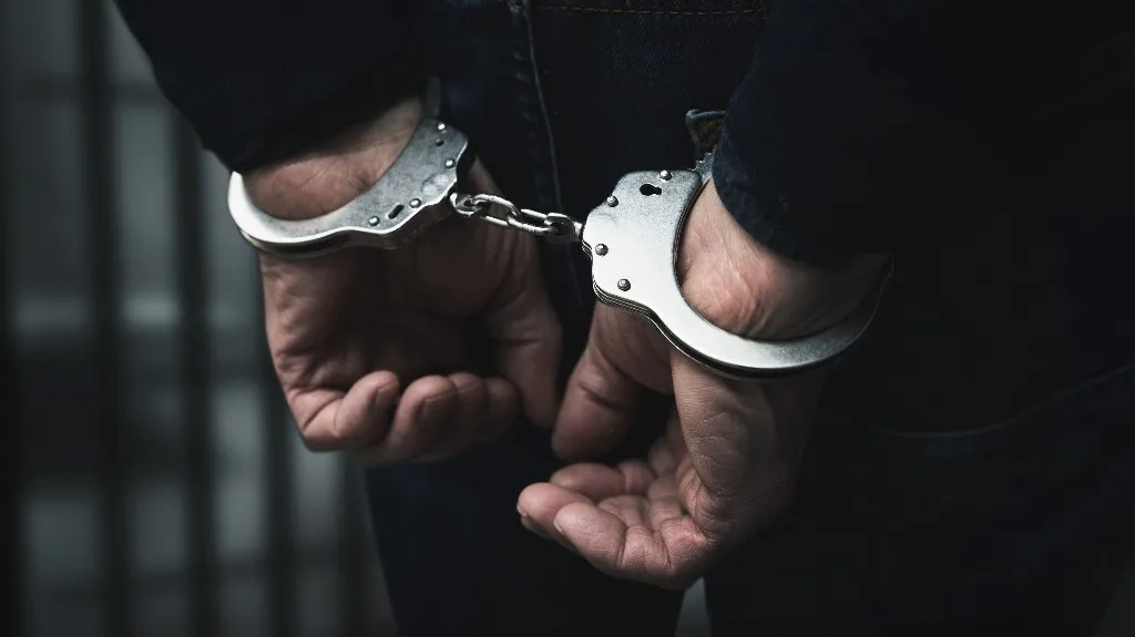 Close-up of a person's hands secured with metal handcuffs behind their back against a dark background.