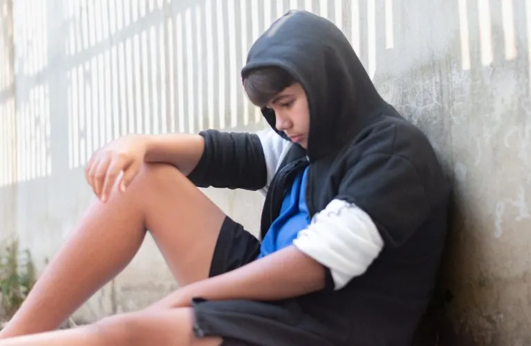 Teenage boy in a black hoodie and blue shirt sitting against a concrete wall, looking down.