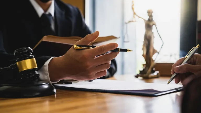 Two professionals in formal attire discussing documents, with a gavel and Lady Justice statue on the desk.