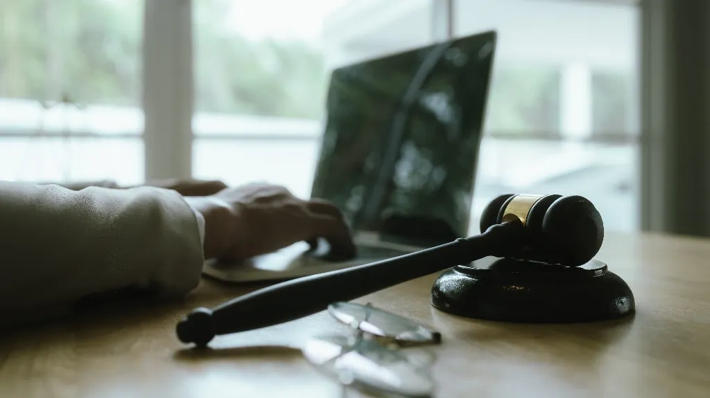 Wooden judge's gavel on desk with eyeglasses and person typing on laptop in background.