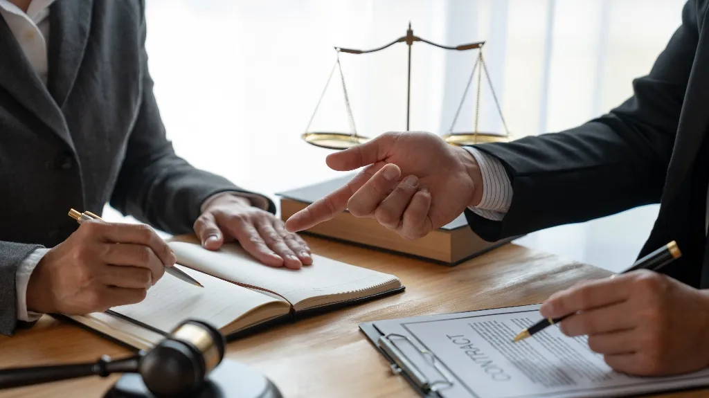 Two professionals in suits discussing a contract with legal scales and a gavel on the desk.