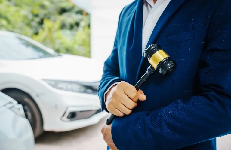 Man in blue suit holding a black and gold gavel, with a white car in the background.