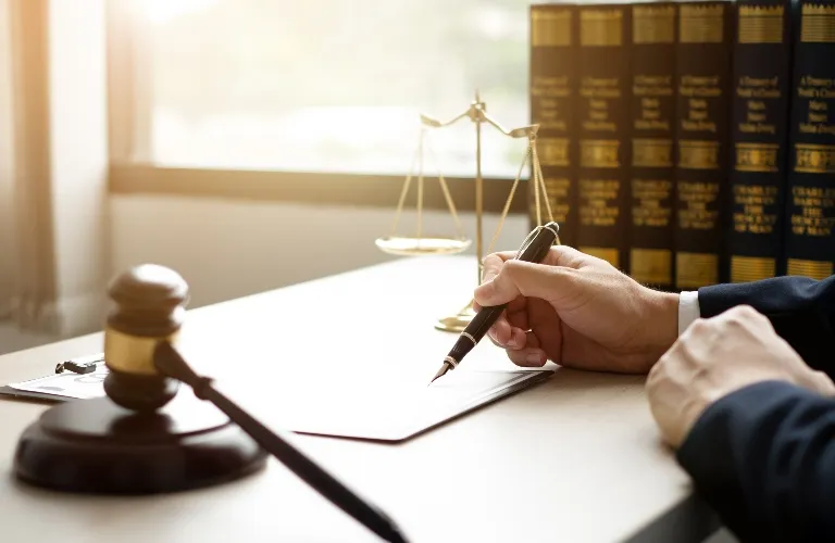 Hand holding pen poised over document on desk with gavel, scales, and law books.