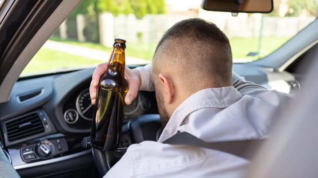 Man holding a dark glass bottle inside a car, leaning on the steering wheel.